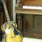 Acoustic guitar standing in front of a vintage upright piano