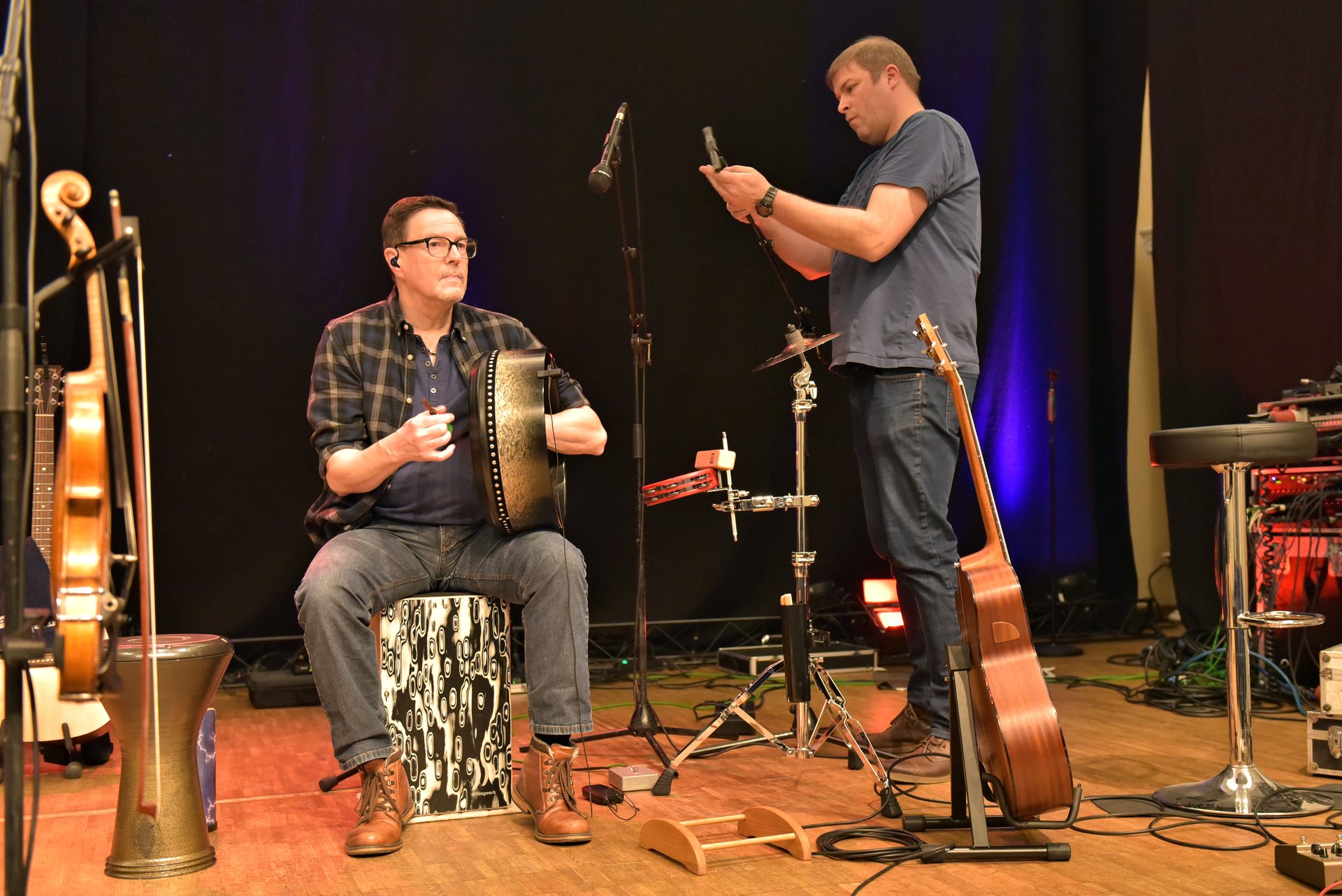 Two musicians setting up during a soundcheck on a small stage, one adjusting a microphone while an acoustic guitar waits on its stand