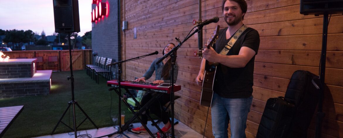 first_gig_guitarist_small_stage How to prepare for your first gig: a smiling guitarist with an acoustic guitar stands at a microphone on a small outdoor venue stage, with a keyboard player, speakers on stands and gig bags beside him