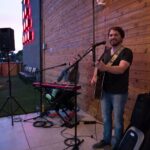 How to prepare for your first gig: a smiling guitarist with an acoustic guitar stands at a microphone on a small outdoor venue stage, with a keyboard player, speakers on stands and gig bags beside him