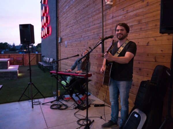 How to prepare for your first gig: a smiling guitarist with an acoustic guitar stands at a microphone on a small outdoor venue stage, with a keyboard player, speakers on stands and gig bags beside him
