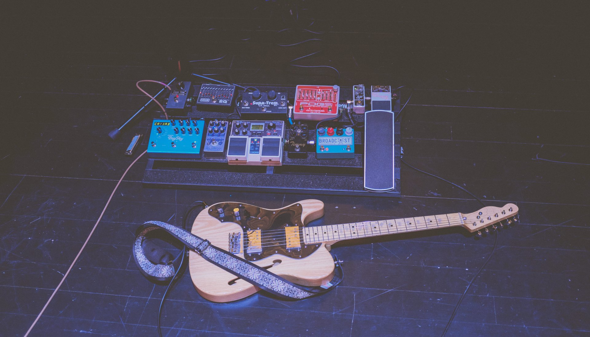An electric guitar with a cream body lying on a dark stage floor below a pedalboard of effects pedals, cabled up and ready for a show