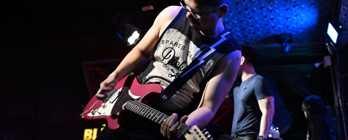 img2_pattaya_solo_guitarist A guitarist playing a red electric guitar on stage under blue lights at a live gig, one side of the guitarist online vs gigs choice