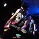 A guitarist playing a red electric guitar on stage under blue lights at a live gig, one side of the guitarist online vs gigs choice