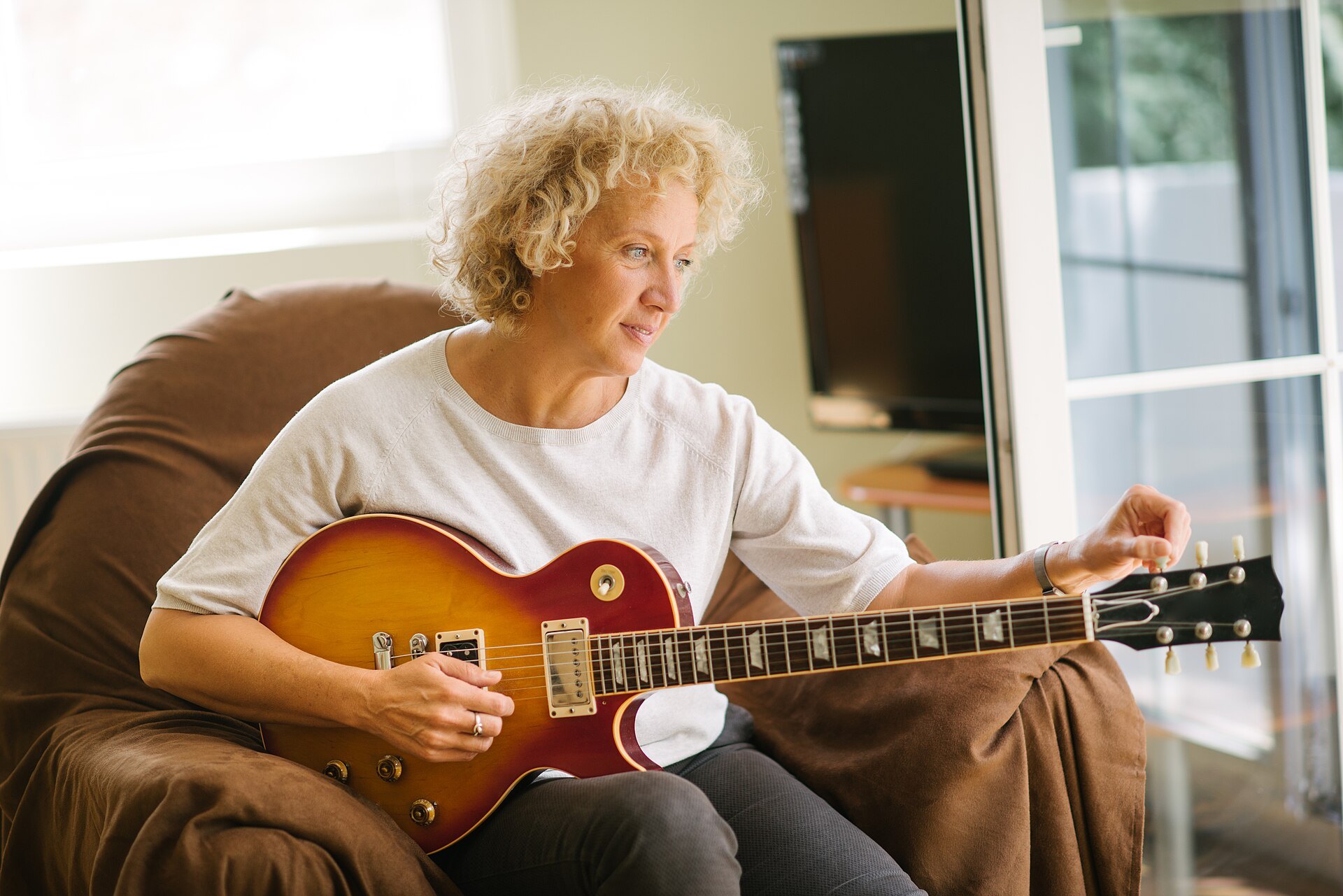 Woman sitting in an armchair at home adjusting the tuning pegs of a sunburst electric guitar